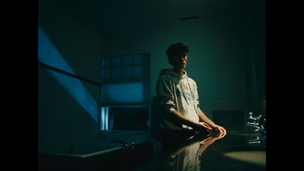 Rhys stands at a sink in the bathroom. Only half of his face is illuminated.