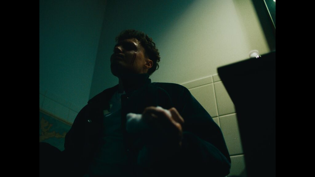 Blaise (played by Garrett Sweere) sits in the half-light of the bathroom. His eye is bleeding and he holds a wet towel.