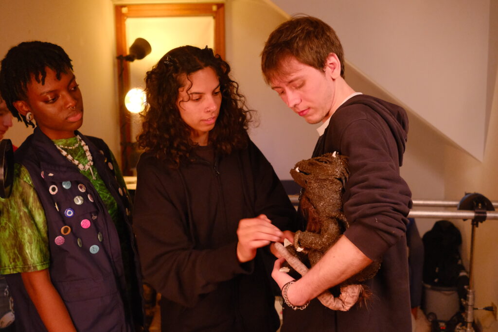 Sasha (production designer) adjusts Miller (puppet) in the arms of Nick (playing Worm Breath) while Aiden (playing Constance) looks on.