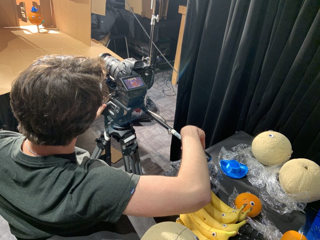 Sam (director) operates a Canon C100 near a table full of googly-eyed bananas, oranges, and cantaloupes.