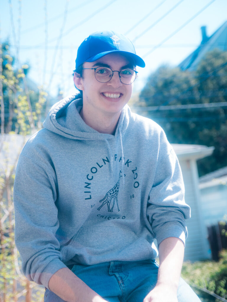 A picture of Samuel Aaberg in a gray hoodie and a blue baseball cap. He's smiling for the camera.