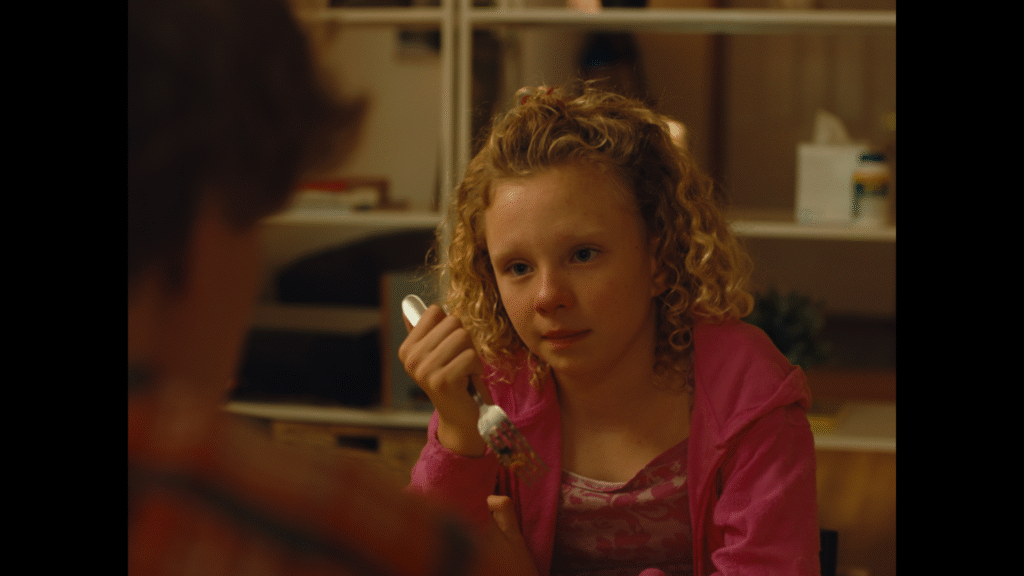 Kayla (an eight-year-old girl), played by Rita Murtagh, holds a fork and looks pensive.