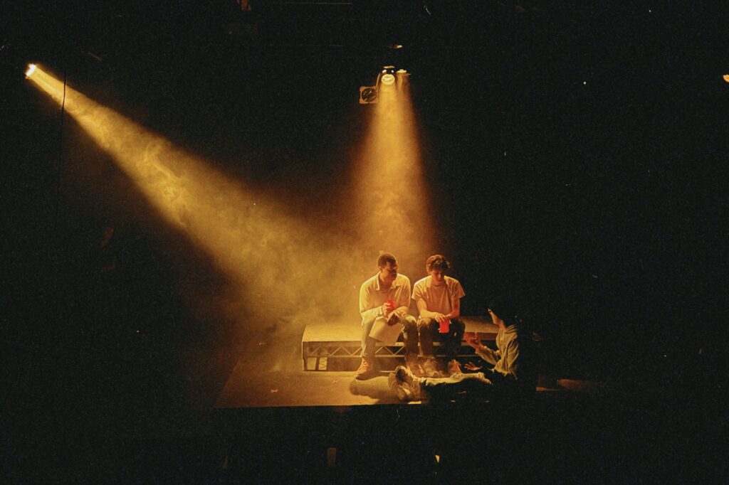 Sam (director) sits on the floor in front of Cliff (playing Trevor) and Quinton (playing Misha) under stage lights.
