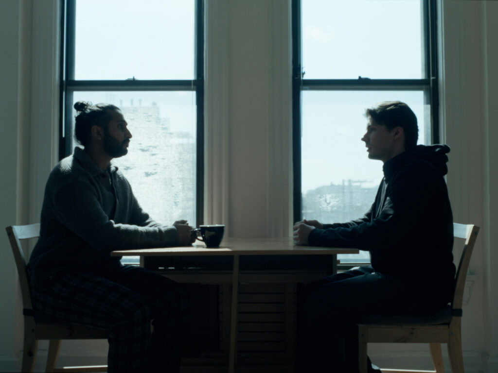 Thatcher and David sit at a table in front of a window. Cold daylight illuminates the scene.