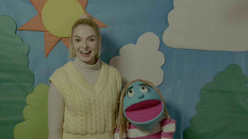 Human Skinface (played by Gabby Seed) and Fuzzy Faelice (puppet, played by Hannah René Strauss) smile in front of a cartoonish paper backdrop.